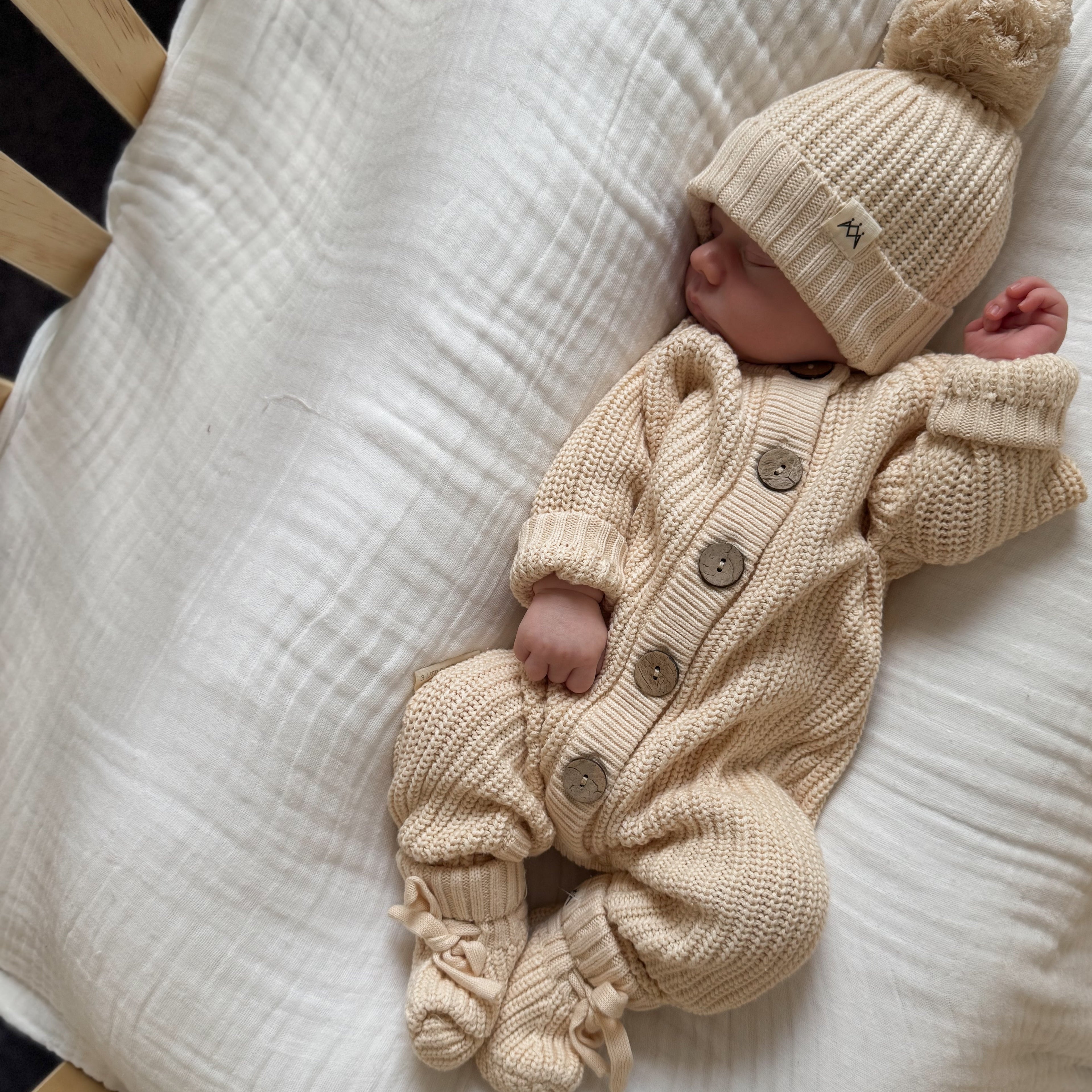 Newborn baby in a beige knitted outfit and hat lying on a white blanket.